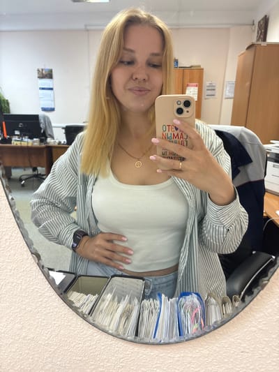 A young blonde woman is taking a mirror selfie in an office environment. She is wearing a light-colored tank top and a vertically striped button-down shirt over it. She has a necklace with a circular pendant and a ring on her right hand. Her phone, with a light-colored case displaying colorful text, is held in her right hand. She is wearing a dark smartwatch on her left wrist and has her left hand on her stomach. The background appears to be an office with desks, chairs, cabinets, and a printer visible in the reflection. The mirror itself has a textured frame and is positioned on a wall with a textured finish. The overall scene suggests a casual moment captured in a workplace. While the city of Заволжье, Russia is mentioned, there are no specific visual cues in the image to confirm this location.