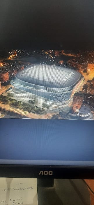 A tilted photograph shows an aerial view of the Santiago Bernabéu Stadium at night, displayed on an AOC monitor. The stadium's redesigned, futuristic exterior is visible, with city lights surrounding it. The monitor's side frame is visible, alongside a handwritten note. The image quality is affected by the monitor's screen, causing vertical lines and distortion.