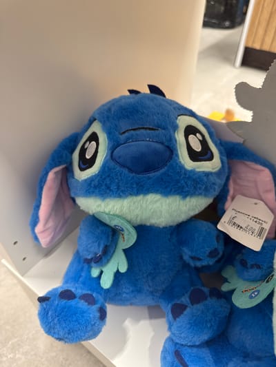 The image shows a close-up, vertically oriented view of a blue plush toy, resembling the character Stitch from Disney's Lilo & Stitch. The toy is lying on a white surface, possibly a shelf or display. The toy's face is prominent, with large black and white eyes and a light blue stripe running down its forehead. It has light pink inner ears and a pink underside to its mouth area. Small light blue leaf-like appendages are visible on its body.

A white tag is attached to the toy's ear. Visible text on the tag includes "Pluszowe zabawki" and a barcode. Below that, the price "8.00" is displayed.

The background is blurred, but hints of a store environment can be inferred, with what appears to be wooden slats and possibly other merchandise in the distance. The lighting suggests an indoor setting, likely during daylight hours. The toy appears to be new and unused, ready for sale. The overall mood is cheerful and inviting, typical of a toy store display.