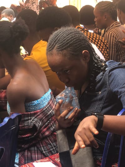 The scene depicts a densely populated indoor or covered gathering at Akpaka Forest Reser, Nigeria. In the foreground, a young female with dark skin, styled cornrow braids, and a denim shirt is observed drinking from a clear plastic water sachet held with both hands. A black smartwatch is visible on her left wrist. To her left, another young female, also with dark skin, is visible from the back, wearing a light blue strapless top with a red and black plaid fabric draped over her waist, and her hair styled in two buns. Both individuals are seated on blue plastic chairs. The background reveals a crowd of numerous individuals, predominantly young adults, seated in rows. Clothing patterns include solid yellow, polka dots, and stripes. Some individuals in the background appear to be interacting with mobile phones. The overall environment suggests an organized assembly or event.