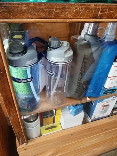 The image captures a close-up view of merchandise displayed within a two-tiered wooden shelving unit, purportedly located in Baza, Myanmar (Burma), though no explicit geographical identifiers are visible. The items are positioned behind a transparent, reflective barrier, likely glass or clear plastic.
On the upper shelf, from left to right:
1. A transparent blue Nalgene water bottle, featuring a grey flip-top lid with a dark grey screw-top and a black strap. Inside, a green mesh filter or infuser is visible, along with a clear straw. The "Nalgene" brand name is prominent in white.
2. A clear transparent Nalgene water bottle, identical in design to the first, with a grey flip-top lid, dark grey screw-top, and black strap. It contains a clear straw and has a white tag attached to its strap. The "Nalgene" brand name is visible in white.
3. Two flexible, collapsible soft water bottles or pouches. The one on the left is dark grey with the word "SPORT" debossed vertically on its surface, and it has a black screw cap. The one on the right is transparent blue, also with "SPORT" debossed vertically, and a black screw cap.
4. In the upper left background, behind the Nalgene bottles, a black box with white text, including "KEYCHAIN LIGHT," is partially visible.
On the lower shelf, arranged beneath the water bottles:
1. On the far left, a silver or light grey cylindrical container, possibly a thermos or insulated mug.
2. In the center, several small boxed items are visible. A prominent yellow box displaying the brand "Brica" is distinguishable. Other white and blue boxes are stacked nearby.
3. On the right, a white box with orange accents is visible, labeled "Camping Light" and featuring an illustration of a lantern. A red and white box is partially obscured above it.
The wooden shelves are light brown, displaying visible scratches and wear, particularly on the horizontal surface separating the two tiers. The overall scene shows various outdoor or travel-related accessories.