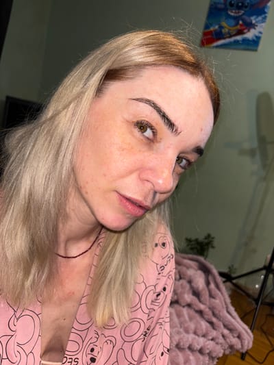 This is a close-up, angled photograph of a woman's face and upper body. She has blonde hair that is styled down, framing her face. Her skin is fair with some freckles and visible blemishes. She is looking off to the side with her eyes open, and her lips are slightly parted. She is wearing a pink garment with a repeated pattern of cartoon characters. A thin, dark beaded necklace is visible around her neck. There are some red marks on her neck and upper chest. In the background, a piece of artwork depicting a cartoon character (possibly Stitch from Lilo & Stitch) can be seen on the wall. The lighting suggests it might be indoors with artificial light.