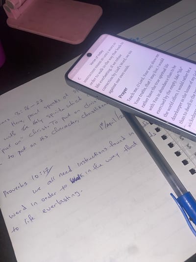 A top-down, close-up shot shows a person's handwritten notes alongside a mobile phone displaying a devotional prayer. The notes, written in blue ink on white paper, include a Bible verse reference (Proverbs 10:17) and the date "1st / April /20". The prayer on the phone discusses walking in God's word and seeking correction. A blue pen rests on top of the paper and the phone, along with a pink phone stand. The scene is set indoors with a dark surface visible in the background.