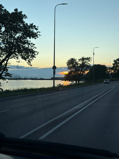 This is a vertical, eye-level, full shot of a road at sunset in Tallinn, Estonia. The sky is a gradient of light blue, yellow, and orange, indicating the sun is low on the horizon. A large tree is silhouetted on the left side of the frame, its branches extending over a body of water. In the distance, a harbor with multiple boats is visible. The road, marked with white lines, curves to the right. A single car drives on the road in the distance. Streetlights are visible along the road. The foreground shows the dark interior of a vehicle, possibly a car or bus, from which the photo was taken.