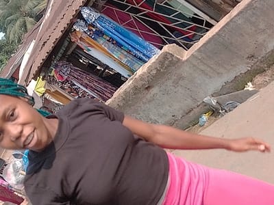 A young woman with teal braided hair, wearing a black short-sleeved top and pink pants, is positioned in the foreground, looking towards the camera with a slight smile or open-mouthed expression. Her left arm is extended to the right side of the frame. The scene is located outdoors in Orie Uratta, Nigeria. In the background to her left, a market stall displays various colorful fabrics and clothing items under a corrugated metal roof. Distant palm trees are visible above the market stall. To her right, a concrete structure, appearing to be a drainage ditch or curb, contains some refuse, including a blue plastic bag and other unidentified debris. The image is captured in bright daylight and has a noticeable tilt.