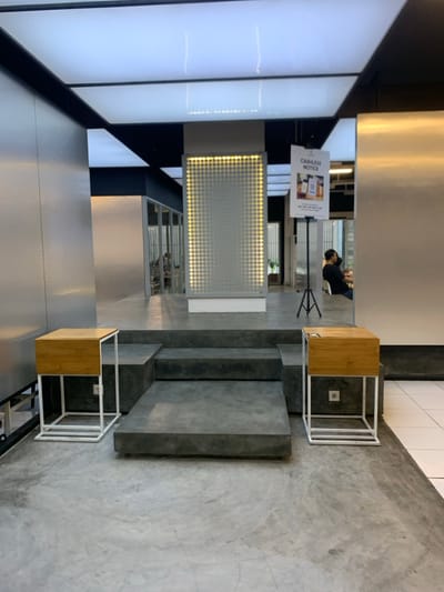 The image depicts a modern interior space with a concrete floor, a set of concrete stairs, and two identical white metal side tables with wooden tops. There is a sign on a tripod stand that reads "Cashless Notice". The sign is situated at the top of the stairs. In the reflection of a large mirror, there is a person sitting at a table. The space appears to be a waiting area or a cafe. The location is the city of Malang, Indonesia, with coordinates (latitude: -7.941, longitude: 112.608).