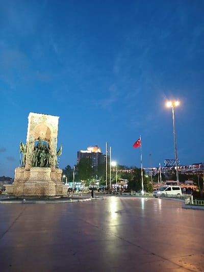 The image shows the Republic Monument in Taksim Square, Istanbul, Türkiye (latitude: 41.037, longitude: 28.985). The monument is a large, rectangular structure made of stone, with a series of statues of people on top of it. The monument is lit up, and there are a few people walking around it. In the background, there are some trees, buildings, a Turkish flag, and a street lamp. The sky is a dark blue, and there are a few clouds in the sky. The square is empty and paved. A white van is parked at the right edge of the square. A tall, thin building with lights on can be seen in the background. The scene is lit by street lights and the lights on the monument. There are a few people standing in front of the monument.