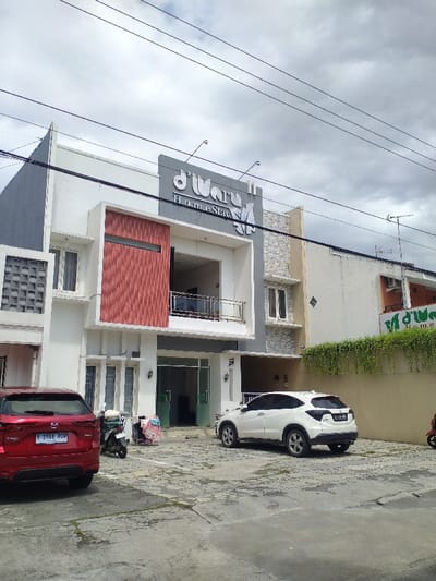 The image shows a two-story building with the name "d'Wara Homestay" on the exterior. The building is located in Surakarta, Indonesia at the coordinates -7.565 latitude and 110.793 longitude. It appears to be a residential building with a parking lot in front. There are two cars parked in the lot, a red car with a license plate and a white car. A motorcycle is parked behind the red car. There is a door and window in the building. The building appears to be newly constructed. The sky is cloudy and there are electrical wires running across the image. There are no people visible. The ground is paved with gray concrete and there is some landscaping on the right side of the image.