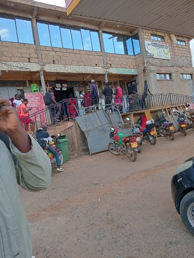 The image shows a scene outside a supermarket in Kipkarren, Kenya. The supermarket is located at 0.607 latitude, 34.964 longitude. The building is made of brick and has large windows. The name of the supermarket is "SUPERMARKET" and it is located in a bustling area with people walking by and motorcycles parked in front of the building.  The scene includes a man taking a picture of the scene with his cell phone.  The man is wearing a green jacket.  There are several other people in the scene.  They are all dressed casually.  The scene is lit by the sun and the sky is blue.