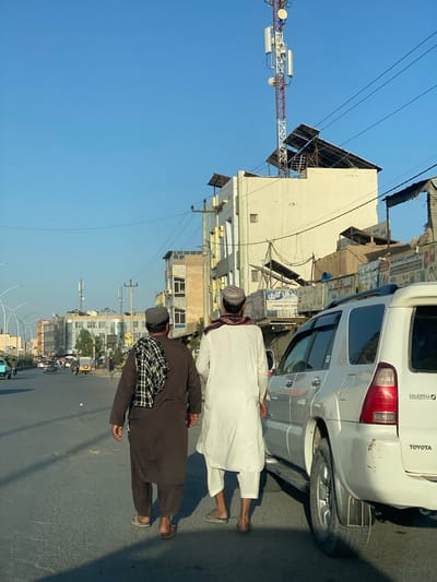 Two men, dressed in traditional Afghan clothing, walk on a street in Kandahar, Afghanistan. The man on the left wears a brown tunic and trousers with a checkered scarf draped over his shoulders. The man on the right wears a white tunic and trousers. A white SUV is parked to their right. In the background, buildings line the street, and a tall communication tower is visible against the blue sky. The street is paved and appears to be a busy area with other vehicles and pedestrians. The overall scene depicts a slice of daily life in the city.