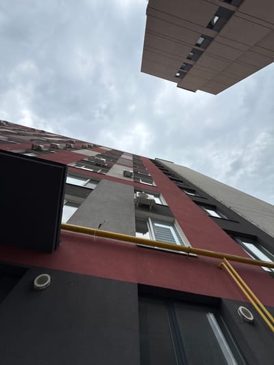 An extreme low-angle, upward-looking photographic view captures two multi-story buildings against an overcast sky in Irpin, Ukraine.
The primary building, occupying the left and center-right of the frame, is a modern residential structure with a facade patterned in alternating horizontal and vertical sections of dark red, light gray, and dark gray/black textured material. Numerous white external air conditioning units are affixed to the facade, predominantly on the red and light gray sections, positioned below windows on various floors. Rectangular windows with white frames are visible, some reflecting the cloudy sky, others showing interior blinds or darkness. A prominent bright yellow gas pipe runs horizontally across the lower dark red section of the facade, then angles upwards and to the right, with smaller yellow pipes branching off. Two circular white louvered vents are observed on the lower dark gray facade.
In the upper right quadrant, a second, distinct building or cantilevered structure is visible. It presents as a large, flat, brownish-beige slab, appearing to hang in the sky from this low vantage point. This structure features horizontal panel lines and several small, dark, recessed rectangular openings or windows.
The sky is uniformly overcast with light gray clouds, providing a neutral backdrop for both architectural subjects. No people or active elements are discernible within the frame.