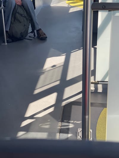 The image captures a close-up view of a person's legs and a backpack inside what appears to be public transportation, possibly a bus or train, in Pilsen, Czechia. The perspective is low, looking upwards towards the ceiling and window area, with a horizontal metal bar dominating the foreground.
The subject's legs are clad in blue jeans and a brown, patterned hiking boot. The foot of the boot is pressed against a dark gray wall, which suggests the person is seated or resting their leg against the interior. To the left and slightly below the legs, a dark green backpack is visible, resting on the floor and partially obscured by the person's clothing.
The floor of the vehicle is a plain dark gray. Sunlight streams in from a window, casting distinct geometric shadows across the floor and wall, indicating it's daytime. The shadows are sharp, suggesting direct sunlight. The interior of the vehicle is utilitarian, with smooth, unadorned surfaces. A yellow strip is visible on the wall towards the upper right of the frame, possibly a safety marking or design element.
The overall mood is one of transit and everyday travel. There are no discernible emotions expressed, as the focus is on the legs and the immediate surroundings. The environment appears clean and well-maintained. The visible text is limited to a small, unreadable label or marking on a silver handle-like object in the upper right quadrant of the image. The time of day is likely midday, given the strength and angle of the sunlight.