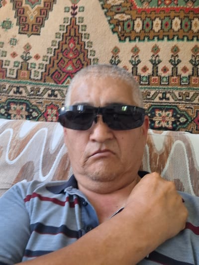 A middle-aged man with short, gray hair and sunglasses is seated on a couch, facing the camera. He is wearing a striped polo shirt. The background consists of a patterned rug with geometric designs. The setting appears to be indoors, likely a living room. There is no identifiable indication of the specific location within Almaty, Kazakhstan.