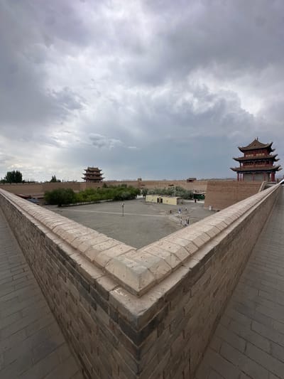 This image depicts a view from the top of a thick, brick-built defensive wall, likely part of the Jiayuguan Great Wall, in Jiayuguan, China. The wall has a stepped coping and forms a sharp V-shaped corner in the foreground, with a walkway paved in bricks extending along its top. Beyond the foreground wall, a vast, flat courtyard with a gravel surface is visible.
Inside the courtyard, several buildings are situated. To the right, a multi-tiered, traditional Chinese pavilion-style structure with a red roof and ornate details stands prominently. Further into the courtyard, another similar, smaller building with a red roof is visible in the distance, to the left of the main pavilion. A line of trees runs along the base of the inner wall, and several low-lying, utilitarian buildings with light-colored walls and dark roofs are scattered throughout the courtyard.
Several individuals, appearing as small figures from this vantage point, are present in the courtyard, walking around. The sky above is overcast with heavy, grey clouds, suggesting a cloudy or potentially rainy day. The overall scene conveys a sense of historical fortification and a subdued atmosphere due to the weather.