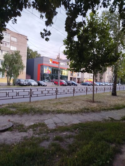 The image depicts an outdoor scene in a city, likely in Chernihiv, Ukraine, showing a street lined with parked cars and a commercial building. In the foreground, a sidewalk and a grassy area are visible, separated from the street by a decorative metal fence. Beyond the fence, several cars are parked along the roadside, including a red hatchback and a dark sedan.
The main structure in the mid-ground is a building with a distinctive facade. It features large glass windows and is accented with a bright orange border. Prominently displayed on the building is the logo and name of a store, reading "SPORT CITY" in large white letters with a stylized orange "S" logo. Inside the store, through the windows, one can see displays with colorful graphics, including what appears to be a sports advertisement with a figure in athletic wear.
Behind the commercial building, apartment buildings and trees form the backdrop. The apartment buildings are made of light-colored brick and have multiple windows. Several trees with green foliage are scattered throughout the scene, with a large tree in the right mid-ground casting a shadow.
The sky above is overcast with a soft, muted light, suggesting it is either early morning or late afternoon. The overall impression is one of a typical urban environment with a focus on retail and residential areas. No people are clearly identifiable as actively engaged in any specific activity, though a figure can be seen walking in the distance near the red car. The atmosphere appears calm and ordinary.