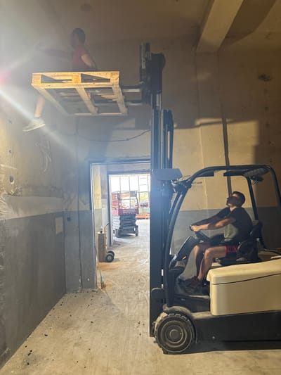 The image shows two men in what appears to be an indoor construction or renovation site. In the foreground, a man in a red t-shirt is partially visible, reaching towards a wooden pallet that is elevated. The lighting is dim, with bright light sources creating a hazy effect, suggesting it might be daytime with artificial lights also in use.
In the background, a man wearing a dark t-shirt and red shorts is operating a forklift. He is gripping the controls and looking downwards, focused on his task. The forklift has a large mast and is likely in the process of moving or positioning materials. The setting appears to be a large, unfinished room with concrete walls and floors. There are exposed pipes and wires, typical of a construction environment. A window or opening in the background reveals daylight and the outline of some equipment, possibly outside the building. The overall atmosphere is one of work and progress within a somewhat rugged setting. The location context indicates Dupnitsa, Bulgaria. No visible text is clearly discernible.