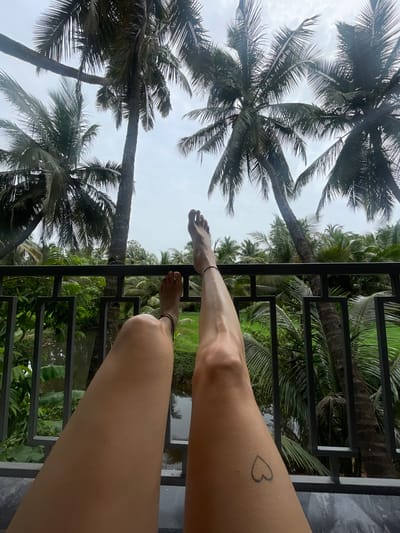An over-the-shoulder view shows a person's legs and feet propped up on a balcony railing. Lush green foliage and tall palm trees dominate the background, suggesting a tropical location. A body of water can be seen through the railing. The person has a small heart tattoo on their left thigh. The scene is likely an outdoor setting, possibly a resort or hotel balcony in Badem, India, given the characteristic tropical vegetation. The overall mood is relaxed and leisurely.