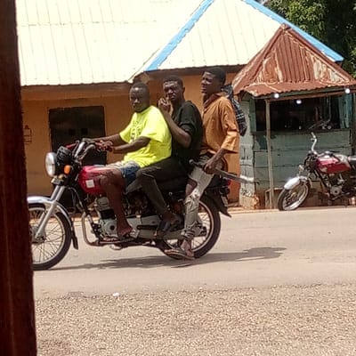 The image captures three young men riding a red and silver motorcycle on a paved road in Mararaba, Nigeria, under bright daylight.

The driver, positioned at the front, wears a bright yellow t-shirt and blue shorts, with flip-flops on his feet. The middle passenger is dressed in a dark t-shirt and dark pants, also wearing flip-flops, and is seen with his hand near his face. The rear passenger, wearing a tan long-sleeved shirt and dark pants, carries a backpack and also wears flip-flops. The motorcycle features a red fuel tank, silver handlebars, and spoked wheels, appearing to be a common model for local transport.

The scene suggests a semi-urban or rural environment. In the background, to the left, is a building with a light-colored corrugated metal roof, possibly white or cream, and blue trim. To the right, a smaller structure, resembling a kiosk or stall, has a distinct rusty brown corrugated metal roof. Green foliage is visible behind this smaller building. Another red motorcycle is parked near the right-side structure. The strong sunlight indicates a clear, sunny day, with visible shadows cast on the road. No legible text is visible on any subjects or objects. The image depicts a common mode of transportation and everyday activity in the region.