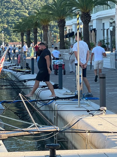 The image depicts a sunny day in Kumbor, Montenegro, with several people visible on a waterfront promenade and near a docked boat. The setting features modern architecture, palm trees, and the water. One man in the foreground is wearing a black t-shirt and shorts and is standing with his left leg extended, appearing to be in motion or posing. Behind him, another man in a white t-shirt and light shorts is walking. Further back, a group of people are gathered, some of whom are dressed in white. The water is calm, reflecting the light. The scene suggests a leisurely atmosphere, possibly a tourist area. There is no visible text in the image.