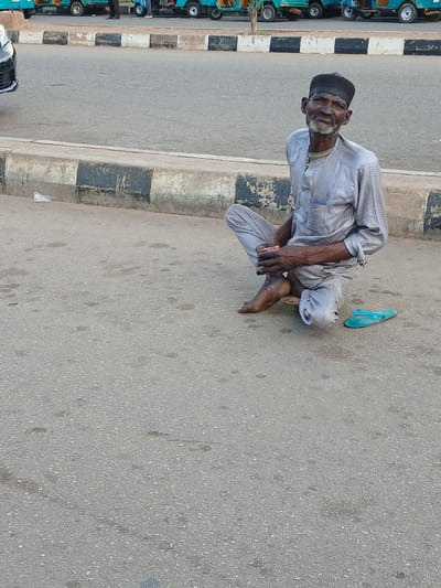 An elderly man, wearing a grey traditional outfit and a black cap, sits on the asphalt next to a curb in Barakin Lerawi, Nigeria. He is looking towards the camera with a slight smile. A single blue flip-flop is visible on the ground to his right. The background shows a paved road with a few parked tricycles.