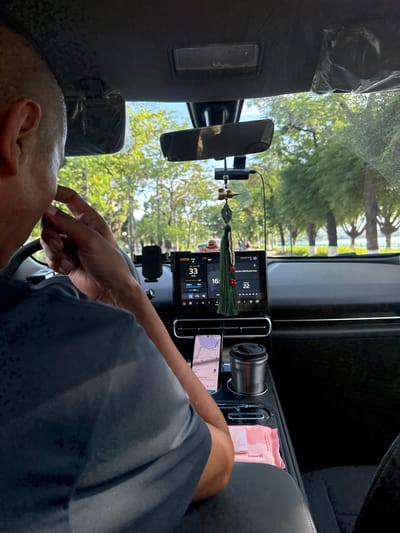 The image shows the interior of a car, likely in Hanoi, Vietnam, as indicated by the text on a wet wipe packet. The scene is viewed from the perspective of a passenger in the back seat, looking towards the dashboard and the driver.
The primary subjects are the driver, whose head and shoulder are visible in the foreground, and the car's dashboard area. The driver is wearing a dark t-shirt and appears to be interacting with something on the dashboard, possibly the screen or a device mounted there. The dashboard features a large touchscreen display showing a navigation map, weather information, and vehicle speed (33 km/h). There are also a car vent, a cup holder with a cup, a phone mounted on a stand displaying a map, and a pack of "gummi" wet wipes. A decorative tassel hangs from the rearview mirror.
The setting appears to be daytime, with natural light filtering in through the windshield, illuminating the trees and greenery visible outside. The weather seems to be clear and warm, judging by the foliage and the presence of the driver's casual attire. The driver's focus seems to be on the dashboard display, and there are no strong emotional cues.
Visible text includes "gummi" on the wet wipe packet and some Vietnamese text on the car's infotainment screen, which appears to relate to navigation and vehicle information. The overall impression is a candid snapshot of a moment inside a car during a drive.
