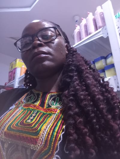In a retail environment, likely within a shop in the city of Howayi, Nigeria, a woman is pictured. She is wearing glasses with black frames and a black garment adorned with colorful embroidery. Her hair is styled in dark, long, curly braids. The background reveals shelves stocked with various products, including bottles and containers that appear to be lotions, shampoos, or similar personal care items. Some of these products are pink and white, while others are in yellow containers with blue lids. The lighting suggests an indoor setting, possibly a beauty supply store or a general store with a personal care section.