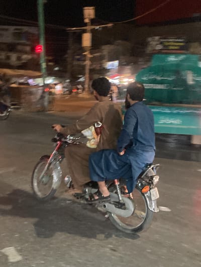 The image depicts a blurred, nighttime street scene in an urban area, identified as Quetta, Pakistan. Two adult males are riding a single motorcycle from left to right across the frame, positioned on a damp or reflective asphalt road surface.
The driver, on the left, wears a brown-colored shalwar kameez or similar traditional garment, with dark hair and a beard. He is seated on the motorcycle's front portion, gripping the handlebars (partially obscured). The passenger, seated directly behind the driver, wears a blue-colored shalwar kameez, also with dark hair and a beard, and has his legs positioned on the motorcycle's rear footpegs. Both individuals and the motorcycle exhibit significant motion blur, suggesting movement during the exposure.
The motorcycle appears to be a standard small-displacement model, with visible chrome accents, a red frame component near the seat, a rear fender, a taillight assembly, and a visible orange rear turn signal on the right side. Both front and rear wheels are visible, with the front wheel particularly blurred.
In the background, on the far left, a red traffic signal light is illuminated, mounted on a tall green pole. To the right of the traffic light, multiple blurred points of light indicate distant streetlights or building illumination. A large, indistinct green vehicle, possibly a truck or bus, occupies the far right side of the background, also heavily blurred. Utility poles and overhead wires are faintly discernible against the dark night sky. The overall impression is a busy street environment during late hours.