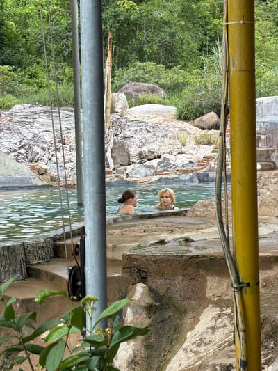 Two young women are bathing in a natural hot spring pool located in a lush, tropical environment. The pool has stone and concrete edges, with steps leading down into the clear, turquoise water. Surrounding the pool are large rocks, dense green foliage, and trees. In the foreground, a thick yellow bamboo pole is visible, along with metal cables and a pulley system. The woman on the left has dark hair pulled back in a bun and is wearing a blue swimsuit. The woman on the right has blonde hair and is wearing a black swimsuit. Both are looking out towards the viewer. The overall impression is a serene and natural setting for relaxation.