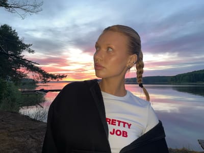 A young, light-skinned woman with blonde hair braided into a ponytail is the primary subject, positioned outdoors near a lake reflecting a vibrant sunset. The woman wears a white t-shirt with the red text "PRETTY JOB" and a black jacket draped over her shoulders. She is looking to the left of the frame. The lake is flanked by trees and vegetation. In the background, a wooden dock and some indistinct structures are visible. The scene is bathed in the soft, colorful light of the sunset. This is likely at the city of Chaykovsky, Russia, given the prompt’s mention of this city.