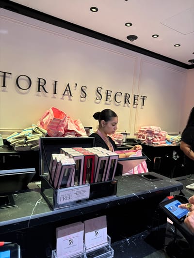 The image shows a retail store with "VICTORIA'S SECRET" branding visible on the wall. A female employee is standing behind a counter, appearing to be attending to merchandise or a customer's needs. Several stacks of folded products, likely apparel or accessories, are visible on the counter. There are also displays of smaller items, possibly perfumes or makeup. A payment terminal is on the counter, with a hand interacting with it, suggesting a transaction is in progress. The environment appears to be an indoor retail setting, possibly a mall or a standalone store. The specific location within New York City is not discernable from the visual information provided.