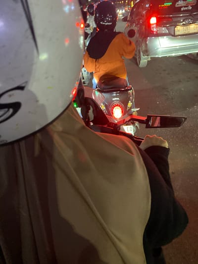 This image shows a person riding a Yamaha motorcycle at night, presumably in Macege, Indonesia. The rider, wearing a white helmet with a black logo and an orange jacket over a black shirt, is seen from behind. To the right, a silver car is visible with its brake lights illuminated, displaying a license plate that reads "DW 1467 EF". The car also has a sticker on the rear window that says "glow.up". In the background, other vehicles and streetlights are faintly visible, indicating a city environment, likely during a traffic jam due to the illuminated brake lights. The motorcycle's taillight is also on, and a portion of its license plate reads "EO". The overall scene depicts typical urban night traffic.