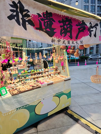 The image shows a street stall in Gongshu District, China, selling a variety of handcrafted items. The stall is covered by a white banner with Chinese characters in black and pink, indicating it is a "new Chinese style cultural and creative" shop. The stall displays numerous small decorative items, including gourds, wooden ornaments, and figurines. There are also baskets and trays filled with these items. A QR code for WeChat Pay is visible on a green stand. The stall is set up on a paved outdoor area, with buildings and a street visible in the background. The lighting suggests it is either early morning or late afternoon. No people are clearly visible in the frame.