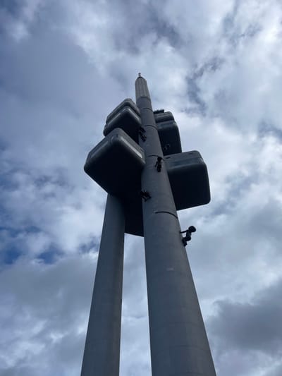 A low-angle view captures the Žižkov Television Tower, located in the Capital City of Prague, Czechia. The tower consists of three substantial grey concrete pillars supporting multiple rectangular prism-shaped modules with rounded edges, which house various facilities. Ten dark, stylized, baby-like sculptures are observed affixed to the tower's vertical pillars, depicting a crawling posture. The uppermost section of the tower culminates in a pointed antenna. The background features a partially cloudy sky with areas of blue and grey clouds.