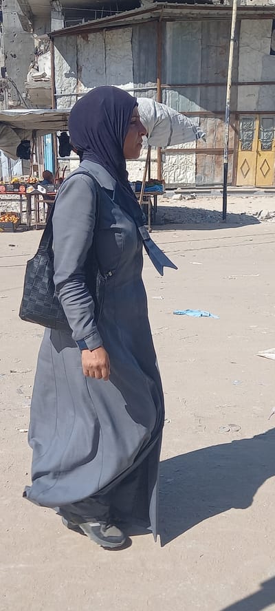 A woman wearing a dark hijab and a long grey dress is walking on a sandy street in Khan Yunis, Palestinian Territories. She has a black woven handbag slung over her left shoulder. In the background, there are damaged buildings and market stalls. The ground is dusty and uneven, with some debris scattered around. The sunlight casts a sharp shadow to the right of the woman.