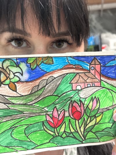 A person with dark hair and brown eyes is holding a colored drawing. The drawing is a stained glass style image featuring a house, flowers, leaves, and water. The colors are primarily red, pink, green, and blue. The location in Zav'yalovo, Russia is not discernible from the image.