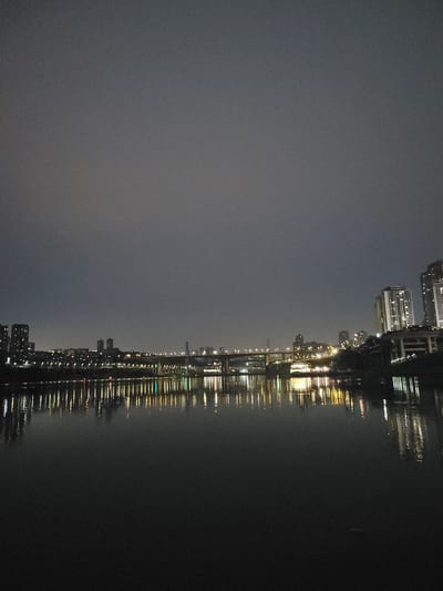 The image depicts a nighttime view of a city skyline reflected in a body of water, possibly a river or bay. The city is in the Shapingba District of China. The left portion of the image shows a dimly lit sky with a dark color, whereas the right has a dark body of water. Reflected in the water is a line of buildings, illuminated by many lights. The reflections are mostly orange and yellow, with occasional green and red highlights. There are bridges and tall buildings visible in the skyline. The water appears to be relatively calm, as the reflections are mostly well-defined.