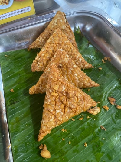 This media file shows a close-up, overhead view of several triangular pieces of fried tempeh arranged on a bright green banana leaf. The tempeh is golden brown and appears crispy, with visible lines indicating it was cut into shape. Small, scattered pieces of fried batter or tempeh are visible on the banana leaf around the main pieces. The banana leaf itself has a glossy sheen, suggesting it might be slightly wet. The tempeh is placed on a stainless steel tray, which takes up the left side of the frame. Part of a yellow sticker with black text is visible on the edge of the tray. The text is partially obscured but includes "Hotline (Chat Only) 0811-2650-705". The scene appears to be a food stall or market setting, likely in North Jakarta, Indonesia, given the visual cues of common food presentation and the style of the text. The lighting is bright and appears to be artificial, possibly from overhead lights in an indoor setting or during the day. There are no people or animals visible in the frame. The focus is solely on the food presentation.