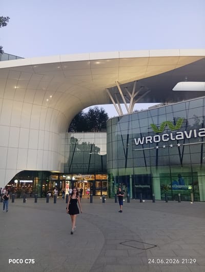 This media file captures a scene outside a modern shopping complex, likely in Wrocław, Poland. The architecture is characterized by a large, curved white overhang and a glass-fronted building adorned with the "WROCLAVIA" logo. Several people are visible in the plaza, including a woman in a black dress walking towards the camera, another person in a black t-shirt and blue shorts standing and looking at their phone, and others in the background near the entrance. One of the shops visible through the glass is Starbucks. The time of day appears to be late afternoon or early evening, with soft, diffused light suggesting fair weather. The overall atmosphere is one of everyday activity and commerce. Visible text includes "POCO C75" and a date and time stamp: "24.06.2026 21:29".