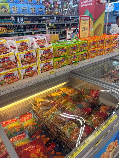 A supermarket aisle displays instant noodles and frozen food in a refrigerated section. Various flavors of "Mie Sedaap" instant noodles are stacked on shelves, including "Goreng" (fried) variants in orange, green, and white packaging. Frozen food items, such as potato products, are visible behind a glass door. Price tags and promotional signs advertise discounts on the products. The layout showcases a typical grocery store setup with categorized items arranged for easy browsing and purchase.
