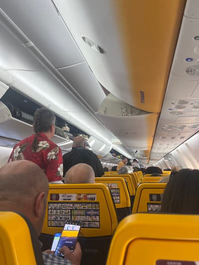 The image depicts the interior of a commercial passenger aircraft, specifically a Ryanair Boeing 737, likely at or departing from Madrid-Barajas Adolfo Suárez Airport, Madrid, Spain. The perspective is from a passenger seat looking forward down the aisle.

In the immediate foreground, the bald head and shoulders of a male passenger are visible. He is wearing a blue and white plaid shirt and is operating two smartphones. His right hand holds a dark-colored smartphone vertically, displaying a brightly colored interface. His left hand holds a lighter-colored smartphone horizontally. In front of him, the back of a yellow aircraft seat is visible, featuring a safety instruction card and a "HOT DRINKS" advertisement with a QR code.

In the mid-ground, a passenger with dark hair styled into a single braid/ponytail is visible from behind, wearing a red shirt with white floral patterns. To their right, another passenger in a dark jacket is partially visible. Further down the aisle, numerous passengers are seated or standing, their backs and heads visible, indicating a full or nearly full aircraft cabin. An "EXIT" sign with a left-pointing arrow is visible above the passengers in the mid-ground.

The aircraft cabin features overhead bins with the "RYANAIR" logo visible on an orange panel on the right side. Some overhead bins are open, revealing luggage stored inside. The cabin ceiling is white and yellow, with integrated lighting and passenger service units. The seats are predominantly yellow with black trim. The overall scene is brightly lit by the interior cabin lighting.