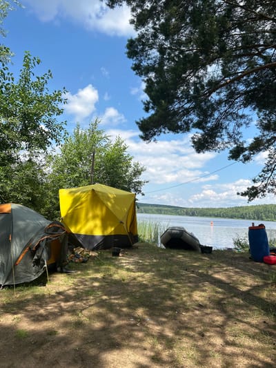 A campsite is set up on the sandy, grassy shore of a large body of water, likely a lake or river, in a wooded area. Two tents are pitched: one is a gray and orange dome tent, and the other is a larger, yellow cube-shaped tent with a visible chimney pipe. In front of the tents, there are logs and some camping gear. An inflatable boat is resting on the bank near the water's edge. To the right, a large blue barrel and a red container are visible. In the distance, across the water, a treed shoreline is visible, and a person can be seen on a paddleboard. The sky is blue with scattered white clouds, and sunlight filters through the branches of a pine tree in the foreground. The location is identified as the city of Chaykovsky, Russia.
