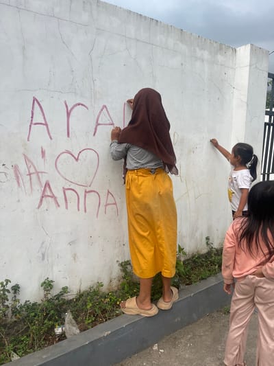 In Desa Batu Layar, Indonesia, three individuals are depicted interacting with a white concrete wall under an overcast sky. The central figure, viewed from behind, is a person wearing a brown head covering, a striped long-sleeved shirt, a mustard yellow long skirt, and tan platform slide sandals. This individual is positioned on a dark grey concrete curb and actively writes on the wall using a red implement, forming the letters "ArAl", a large heart symbol, and the name "ANNA". To the right of the central figure, a smaller child with dark hair in a ponytail, wearing a white t-shirt, also marks the wall with a lighter-colored implement. A third child, partially visible on the far right, wears a light pink long-sleeved top and light pink pants. The white wall exhibits surface cracks and discoloration. Below the wall, a narrow strip of ground contains sparse green vegetation and a discarded plastic bag. A black metal gate structure is partially visible in the background on the right.