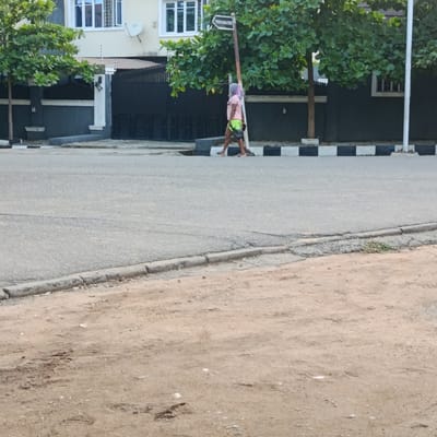 In Gwarinpa, Nigeria, a lone individual walks across a paved road. They are wearing a light-colored t-shirt and bright green shorts. The person's head is covered with a light pink garment, possibly a scarf or veil. They are carrying a dark object in their right hand. Behind the individual, a street sign with white lettering on a black background is attached to a pole. The background features a dark gray building with white-trimmed windows and a black metal gate. Lush green trees with broad leaves are positioned in front of the building. The road surface is asphalt, and a concrete curb with black and white checkered pattern separates the road from the sidewalk. The foreground of the image shows a dirt area with some tire tracks.