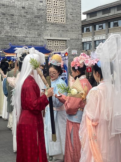 The image depicts a group of individuals in traditional Chinese Hanfu attire gathered outdoors in Shapingba District, China. In the foreground, a person with long white hair, wearing a red patterned robe, holds a black-hilted sword-like object vertically, adorned with green foliage near the top. To their right, a woman with glasses, dressed in a light blue Hanfu with a large gold-colored headpiece accented by blue flowers, observes the sword-holder. Adjacent to her, a woman in a peach-colored Hanfu adorned with a large pink and white floral headpiece holds a bouquet of white flowers wrapped in paper featuring Chinese characters. Partially visible on the far right is another individual in a sheer light pink Hanfu with a white veil headpiece. In the background, a grey brick building with patterned square vents is visible on the left, and a multi-story building with traditional Chinese architectural elements and balconies is on the right. A blue banner with Chinese characters, a hot pot icon, and a Pepsi logo is present behind the subjects, along with a red directional arrow sign pointing right. The ground is an undifferentiated urban surface.