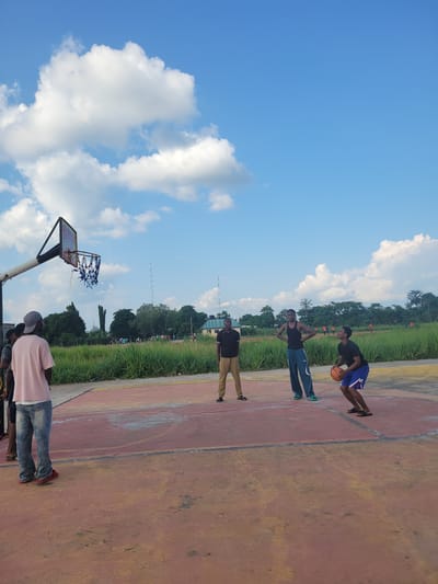 The image shows four people on an outdoor basketball court. The court is made of a reddish-orange material, with yellow and white markings. There is a basketball hoop and backboard on the left side of the frame. Tall green grass borders the court, and beyond that, trees and some buildings are visible. The sky is a bright blue with scattered white clouds, suggesting it is daytime and the weather is pleasant.
One person, wearing a black t-shirt and blue shorts, is in mid-action, holding a basketball and appearing to dribble or prepare to shoot. Another person, in a black tank top and blue pants, stands with hands on hips. A third person, in a black shirt and khaki pants, stands further down the court. The fourth person, wearing a light pink shirt and jeans, is partially visible in the foreground. Their poses suggest they are engaged in a casual game or practice session. The overall mood appears relaxed and active, typical of an outdoor recreational setting. No visible text is present in the image. The context provided indicates the location is Egbagu, Nigeria.