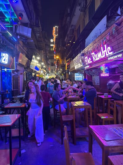 This image depicts a bustling street scene in Hanoi, Vietnam, at night. The street is lined with businesses illuminated by vibrant neon signs and string lights. Several people are present, some walking, while others are seated at tables outside establishments.
One prominent establishment is a bar or club named "The Rumble," indicated by a large, brightly lit sign. Flags of various countries are strung across the front of this venue. Nearby, a sign for "Free Karaoke Sport-Dance" suggests entertainment options. Another sign with the number "2B" is also visible.
The environment is characteristic of a busy urban street at night, with a mix of buildings, signs, and people. The overall atmosphere appears lively and social, with people enjoying the evening outdoors. The ground is paved, and the scene is dominated by artificial lighting, creating a colorful and somewhat chaotic ambiance typical of a nightlife district.