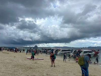 A crowd of people are gathered on a sandy beach in Tambon Phru Nai, Thailand, under a heavily overcast sky. Numerous speedboats are moored in the shallow water near the shore, with some people boarding or disembarking from them. The beach is lined with more speedboats, and in the background, distant islands are visible through the hazy atmosphere. People are dressed in casual beach attire, and some are carrying bags or wearing backpacks. The overall scene suggests a popular tourist destination with active boat tours.