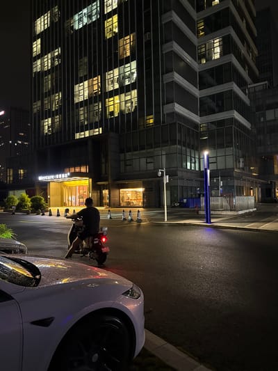 An exterior, ground-level, medium shot captures a nighttime scene in the Huangpu District, China, outside the Mercure hotel. A person, seen from behind, is seated on a scooter. The scooter is parked on a wet, dark asphalt road, with a white car to the left also appearing wet and reflecting the building's lights. In the background, a multi-story building displays numerous illuminated windows, suggesting it is occupied or actively lit from within. A blue illuminated light fixture stands near the center of the frame on the sidewalk. The scene is characterized by artificial lighting from the building and the street lamp.