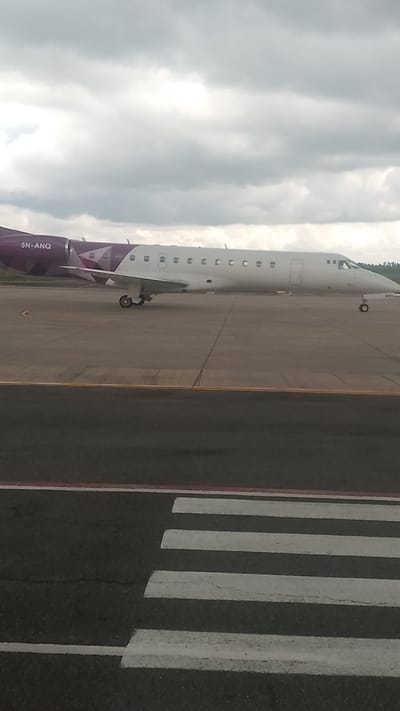An Embraer ERJ-145LR aircraft, registration 5N-ANQ, is parked on the tarmac at Orerokpe Airport in Orerokpe, Nigeria. The aircraft has a white fuselage with purple accents on the tail and wingtips, featuring a geometric design. The registration number "5N-ANQ" is visible on the rear fuselage. The plane is positioned on a gray asphalt surface, with visible runway markings, including a pedestrian crossing in the foreground. The sky is overcast with gray clouds.