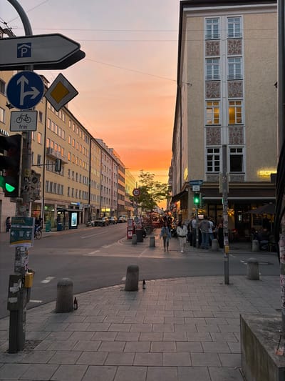 The image depicts an outdoor urban scene in Munich, Germany, likely during sunset given the warm orange and pink hues in the sky. The perspective is from a sidewalk looking down a street lined with buildings.
On the left side of the image, a traffic light is visible, showing a green signal. A lamppost next to it is adorned with various signs and stickers, including a parking sign with a 'P', a directional sign indicating a left turn and straight path, a diamond-shaped warning sign, and a sign with a bicycle symbol and the word "frei".
The buildings along the street are of varying architectural styles. On the left, there are multi-story buildings with light-colored facades, some with balconies. On the right, a prominent building has a cream-colored facade with decorative floral patterns between the windows. Some windows on this building are illuminated from within.
In the street, traffic is present, with cars and a motorcycle visible. Pedestrians are walking on the sidewalk. Some individuals are gathered outside a building on the right that appears to be a cafe or restaurant, with tables and umbrellas set up.
The sidewalk is paved with rectangular stone tiles. Several cylindrical stone bollards are placed along the edge of the sidewalk. Two brown glass bottles are visible on the sidewalk, one near a bollard and another further down the street.
In the background, the sky is a prominent feature, filled with the vibrant colors of sunset. Trees are also visible, adding to the urban greenery. The overall impression is a bustling city street transitioning into the evening.